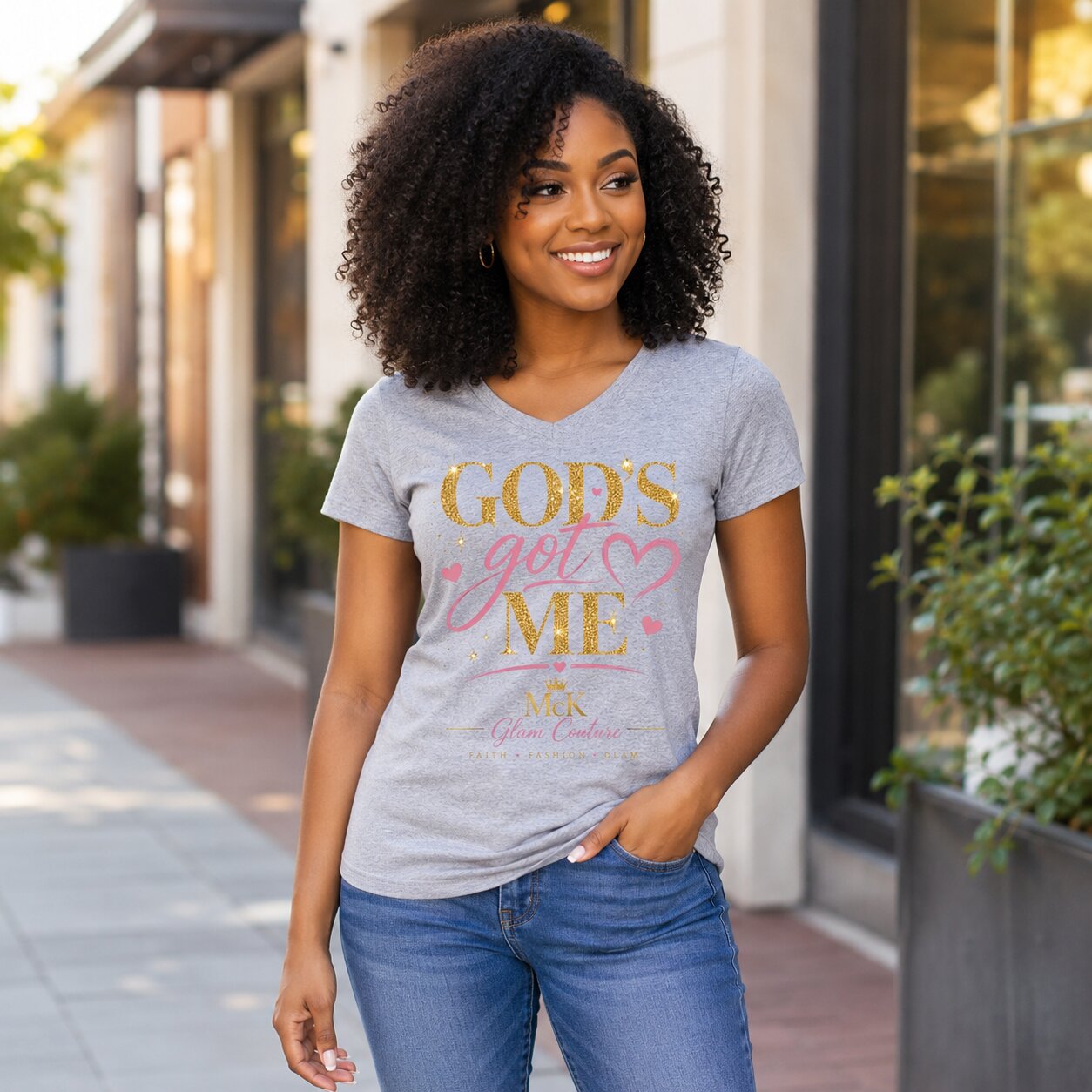 A woman with curly black hair stands outdoors wearing a heather gray V-neck t-shirt with a gold and pink graphic that reads 'GOD'S get ME' and blue jeans.