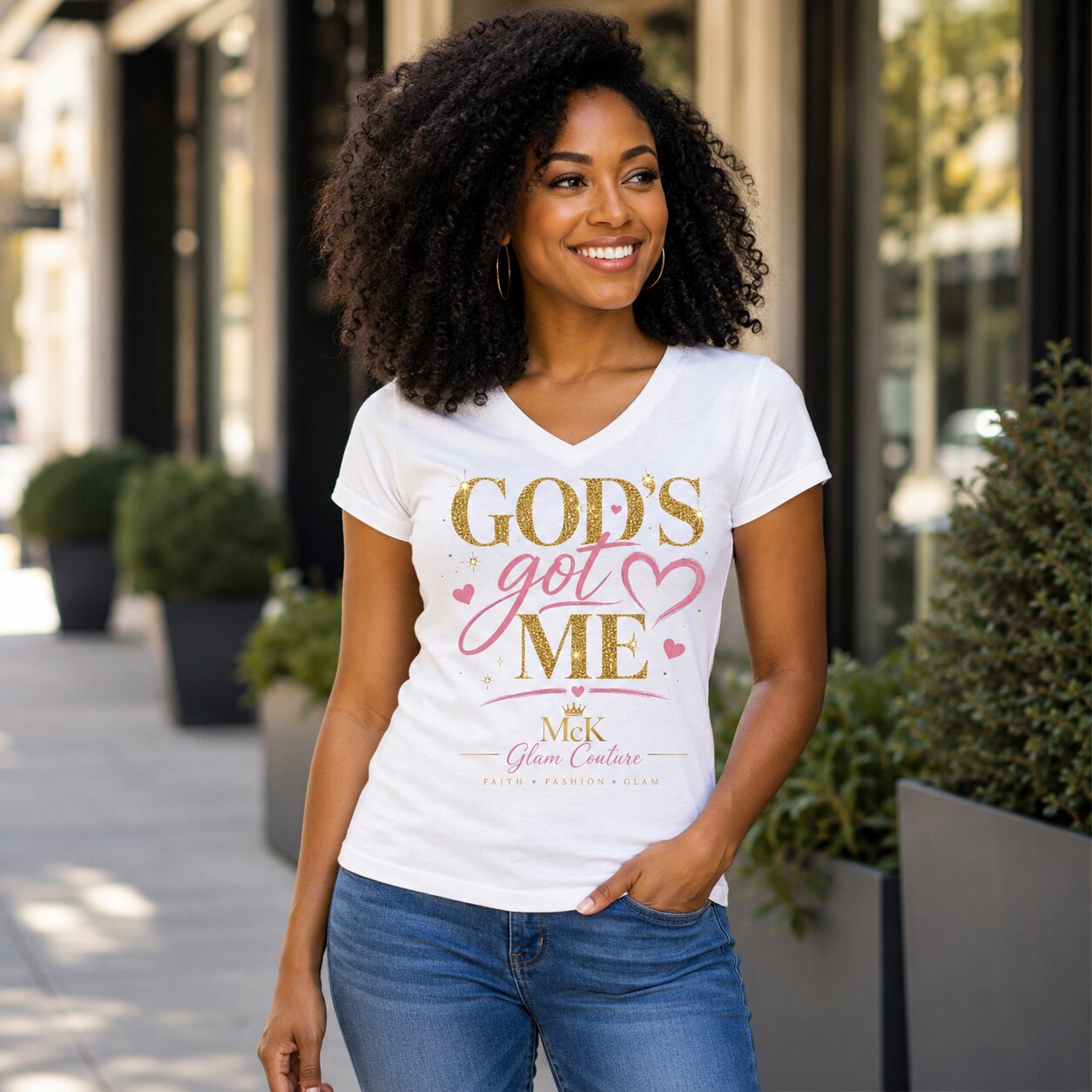 A woman wearing a white graphic t-shirt with 'GOD'S got ME' printed in gold and pink lettering, standing outdoors on a city sidewalk.